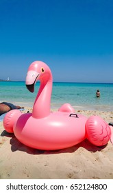 Giant Inflatable Pink Flamingo Pool Float Toy On The Sunny Beach With White Sand And Crystal Clear Blue Sea Water 