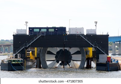 Giant tidal turbine docked in a harbor. It is held in a ship designed to hold and transport it. Sky is overcast. Walkways on ship give sense of scale. Identifying marks removed. Room for text.