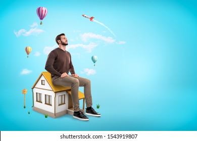 Giant Young Man Sitting On Roof Of Small House Like On Chair, Looking Up In Blue Sky With Copy Space And Dreaming. Thinking About Future. Private Property. Building Market.