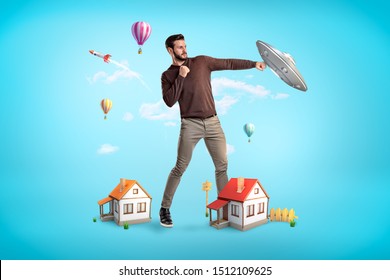 Giant young man with two small houses at his feet, standing in half-turn and fighting a UFO. Protect your home. Develop security systems. Your home is your castle.