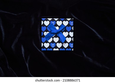 Gift Box With The Image Of Blue Hearts For Valentine's Day On A Black Velvet Background. Valentine's Day Gift For Him. Bright Strong Box In The Center Of The Photo On A Dark Background