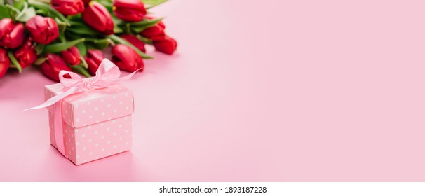 Gift Box With A Pink Ribbon In White Polka Dots On A Background Of A Bouquet Of Red Tulips With Copy Space For Text. Wallpaper Or Banner For Gift Shop, Flower Shop Or Jewelry Store. Valentine Day.