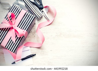 Gift box wrapped in black and white striped paper with pink ribbon and wrapping materials on a white wood old background. Vintage style.