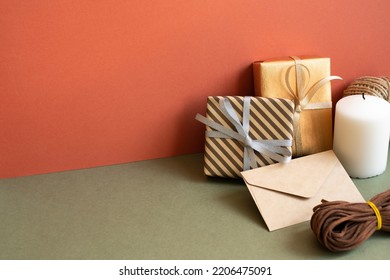 Gift Box And Wrapping Roll, Envelope, Candle On Khaki Table. Red Wall Background. Anniversary, Birthday, Christmas, Greeting Concept