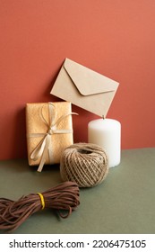 Gift Box And Wrapping Roll, Envelope, Candle On Khaki Table. Red Wall Background. Anniversary, Birthday, Christmas, Greeting Concept