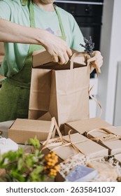 Gift boxes with handmade soaps are prepared on a kitchen table while a person ties a kraft bag. DIY business visuals, handmade product packaging, entrepreneur life, craft fair prep