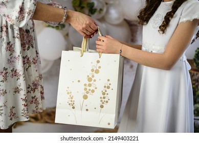 A Gift From Godparents For The First Holy Communion. A Woman Giving The Girl A First Communion Gift. First Communion Gifts In Decorative Bags.