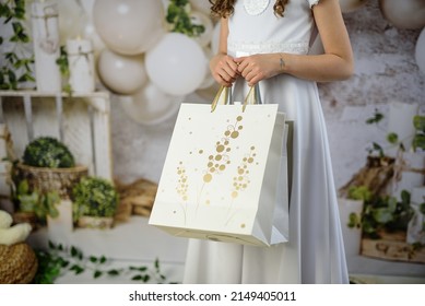 A Gift From Godparents For The First Holy Communion. A Girl In A White Communion Dress With Gifts