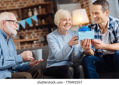 Gift With Love. Cheerful Young Man Giving A Birthday Present To His Happy Elderly Mother While His Father Sitting Next To Them And Drinking Coffee