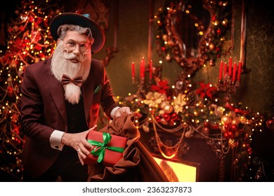 Gift from stylish Santa Claus. Handsome modern Santa Claus in an elegant festive costume gives a gift and smiles joyfully while standing in a magical festive setting at home. Christmas and New Year. - Powered by Shutterstock