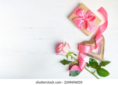 Gift, wooden heart, pink rose, polka dots ribbon on wooden white background. Top view, copy space.