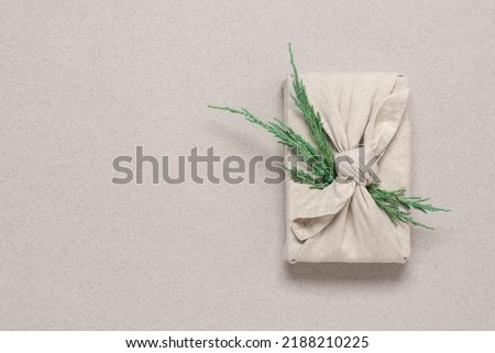 Gift wrapped in fabric with green juniper branch, beige craft paper background. Christmas or New Year's gift in furoshiki style. Zero waste concept. Top view, flat lay, selective focus.