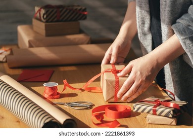 Gift wrapping in eco packaging. Young woman making bow on kraft gift box with red thread on a wooden table. Packaging and preparation of gifts for the celebration.