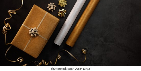 Gift Wrapping Service. Wrapped Golden Present Box With Wrapping Paper Rolls On Dark Stone Background. Banner Copy Space