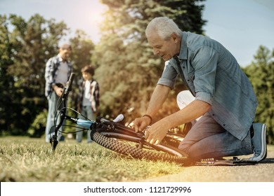 Gifted Fingers. Loving Handy Grey-haired Man Repairing A Bicycle While A Boy And His Dad Standing And Talking