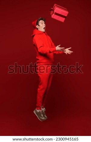 Gifts. Cute teen boy in red hoodie and a red hat catches a gift box and looks with surprise. Studio full-length portrait on a festive red background. Christmas and New Year celebration. 