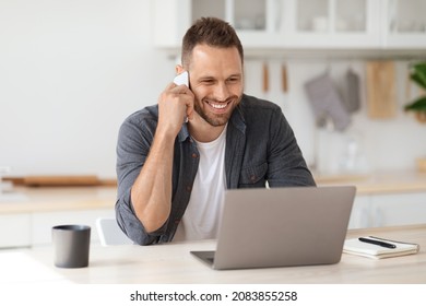 Gig economy concept. Young positive man freelancer working remotely at home, looking at laptop and consulting client via cellphone, sitting at kitchen - Powered by Shutterstock