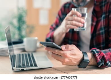 Gig Worker Drinking Water And Using Mobile Phone In Home Office During Freelance Project Work