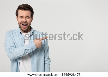 Giggling millennial man wearing blue shirt points finger at empty copy space for text posing isolated on grey studio background show laughing at something shameful awkward and strange, mockery concept