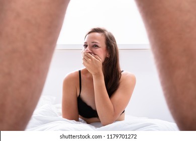 Giggling Young Woman Sitting On Bed Looking At Male's Genital