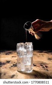 Gin And Tonic Cocktail Recipe, Pouring Gin From A Jigger Into A Highball Glass Filled With Ice, Wooden Table, Dark Background