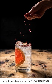 Gin And Tonic Cocktail Recipe, Sprinkling A Drink With Pink Peppercorn, Highball Glass, Wooden Table, Dark Background