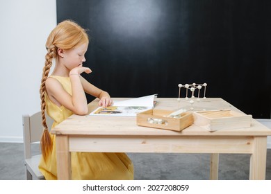 Ginger Girl With Long Braids Sitting Behind A Table, Looking At A Coloring Book. At A Child Learning Center.