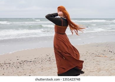 Ginger girl in viking dress at the beach of the Baltic sea. Redhead viking woman on the beach