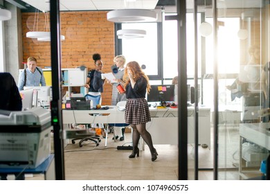 ginger secretary in casual clothers standing with tea and cell phone in the center of busy room - Shutterstock ID 1074960575