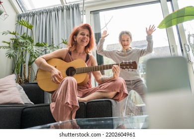 Ginger Tattooed Woman Playing Guitar While Her Child Daughter Dancing Nearby And Recording Video For Tik Tok. Family Concept 