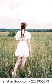 Ginger young woman in field, rear view. Flowers in pigtails. White dress and white sky, copy space.