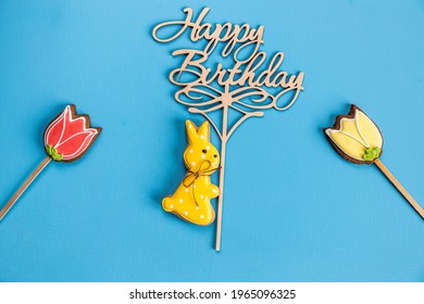 Gingerbread Bunny Holding A Happy Birthday Sign And Two Flowers On Blue Background