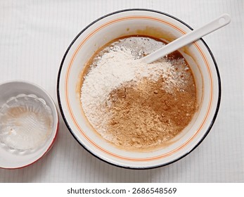 Gingerbread cake baking process: dry ingredients, flower and spices, added over wet ingredients. Large white enamel mixing bowl with red rim, white plastic spatula, smaller empty bows next to it.