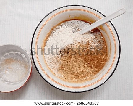 Gingerbread cake baking process: dry ingredients, flower and spices, added over wet ingredients. Large white enamel mixing bowl with red rim, white plastic spatula, smaller empty bows next to it.