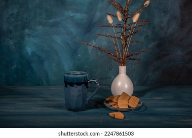 Gingerbread cookies on a saucer, a cup of tea and a bouquet of dry branches in a vase on a dark blue background. - Shutterstock ID 2247861503