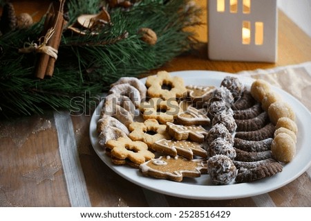 Gingerbread, Linzer and other types of Christmas cookies on a white plate, a festive tablecloth, an Advent wreath and a white decorative house with a burning candle in the background. Selective focus
