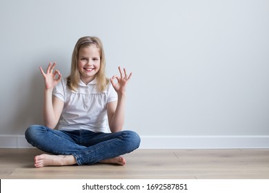 Girl 10 years old sitting on the floor in the room. Barefoot. Looks at the frame, smiles. Shows a gesture that everything is fine .Mockup. Grey background.