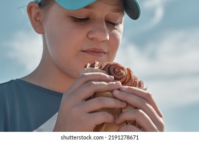 A Girl 10-12 Years Old In A Cap Eats Pastries Against The Background Of A Blue Sky On A Summer Day Outside. Teenager Eating A Cinnamon Bun, Close-up.