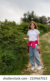 A Girl Of 11 Years Old With Red Hair In Blue Jeans, A White T-shirt And A Pink Jacket Stands Near A Bush In An Olive Garden.