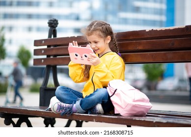 A girl of 6-7 years old holding tablet computer in her hands. She sits outside on a bench. Advertising new gadget, educational app. Games. Children use technology.