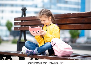 A girl of 6-7 years old holding tablet computer in her hands. She sits outside on a bench. Advertising new gadget, educational app. Games. Children use technology.