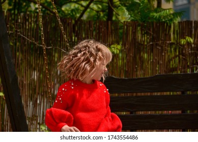 Girl 7 Years Old In Red Clothes On The Street On A Swing. Little Curly Blonde Girl On A Swing In Spring. Schoolgirl. 1st Grade Student. Spring, Nature, Childhood