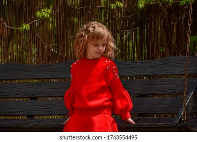 Girl 7 Years Old In Red Clothes On The Street On A Swing. Little Curly Blonde Girl On A Swing In Spring. Schoolgirl. 1st Grade Student. Spring, Nature, Childhood