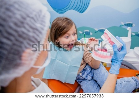 A girl of 7-8 years old looks in surprise at the dummy of the dental jaw, which is told to her by a woman dentist in a dental clinic.