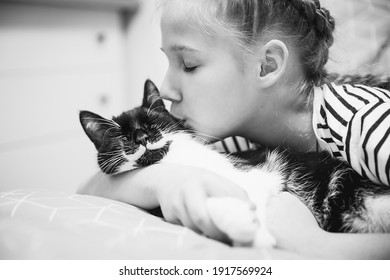 A girl of 8-9 years old hugs and kisses a beautiful black cat with a white mustache, which has closed its eyes with pleasure and smiles. Love for cats, taking care of pets. Pet Day, World Cat Day. 