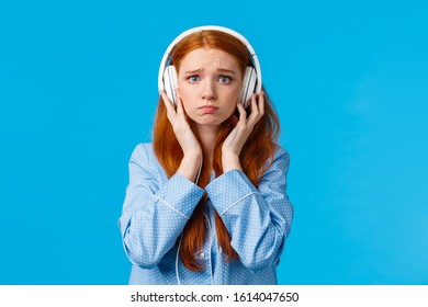 Girl About Cry From Emotional Song. Upset Redhead Girlfriend Suffering Breakup, Sobbing Listening Music In Large White Headphones, Standing Blue Background In Nightwear, Unhappy