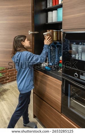 Girl adding container of peanuts to 72 hours survival kit stored in cupboard. Female preparing emergency food pantry in kitchen for disasters. Alert, foresight and self sufficiency strategy.