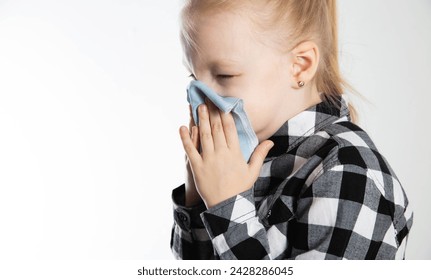A girl aged seven years old in a checkered shirt blows her nose into a handkerchief on a white background. The concept of nasal congestion due to allergies and acute respiratory infections. 