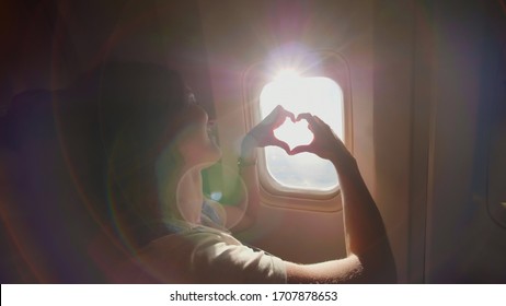 The girl at the airplane's porthole makes a heart out of her hands.