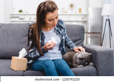 girl with allergy holding facial tissue and stroking cat on couch